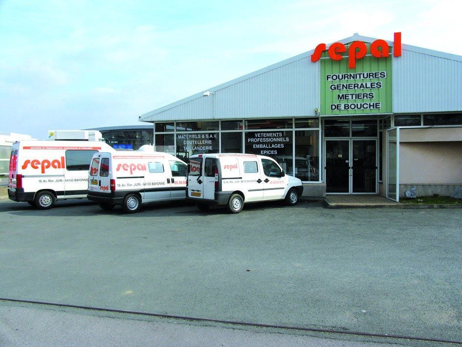 Un bâtiment commercial blanc avec l'inscription Sepal en lettres rouges, devant lequel trois camionnettes blanches sont garées sur un parking pavé.