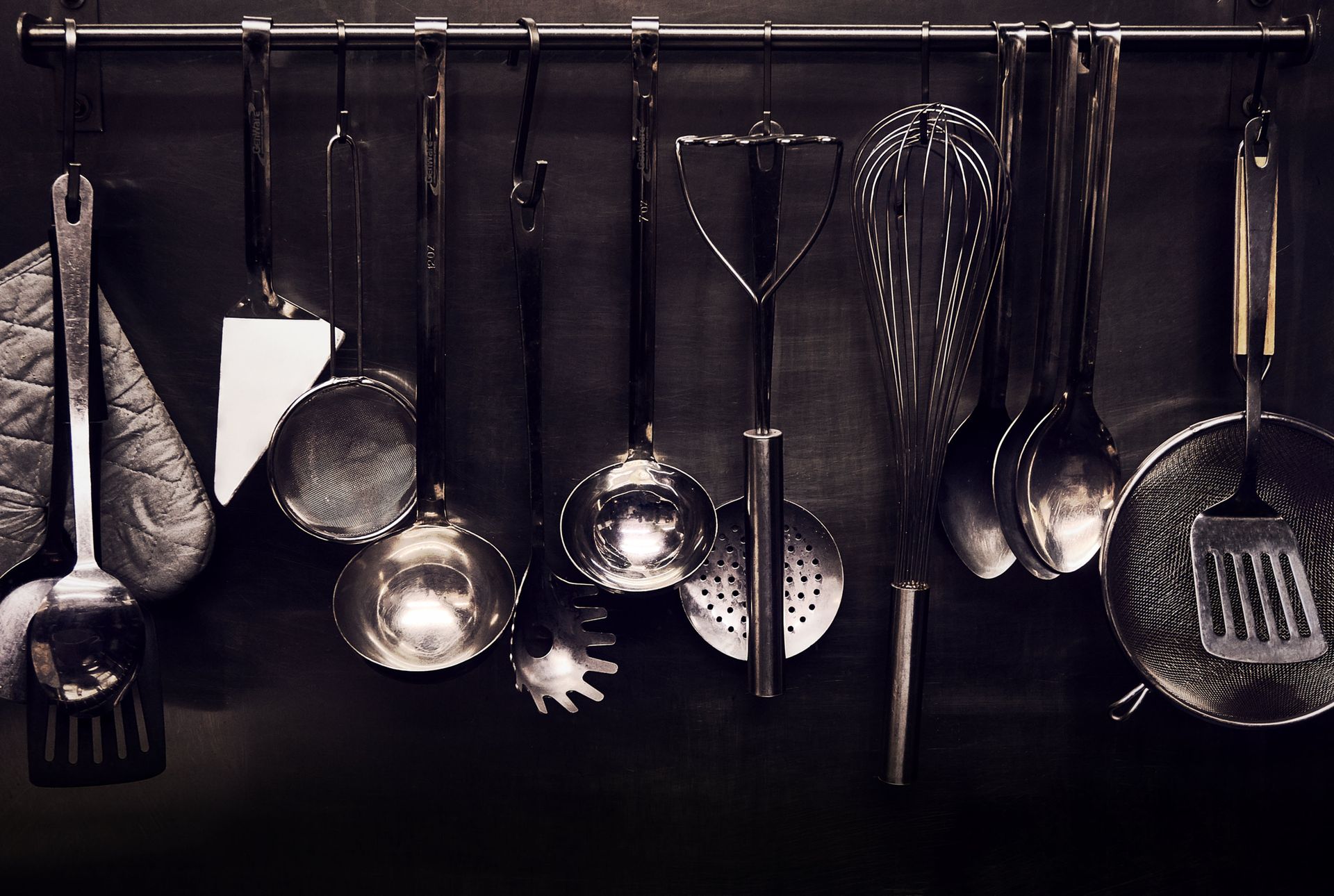 Une collection d'ustensiles de cuisine en argent, comprenant un fouet, une louche et une spatule, est suspendue à un support contre un mur noir.