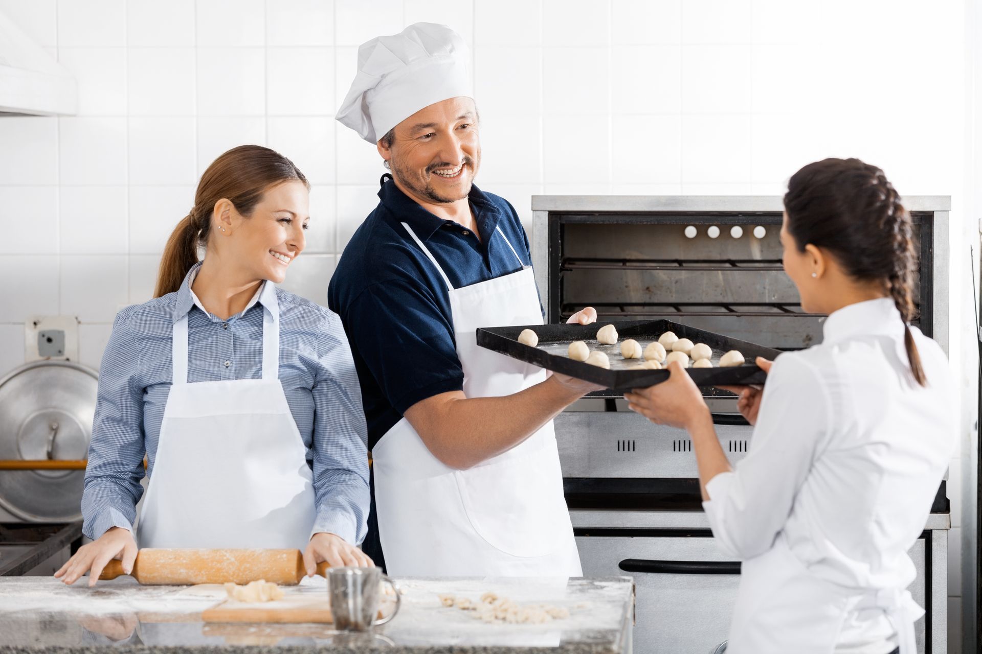 Dans une cuisine professionnelle, un boulanger, une femme et un étudiant préparent une plaque de petits pains à enfourner.