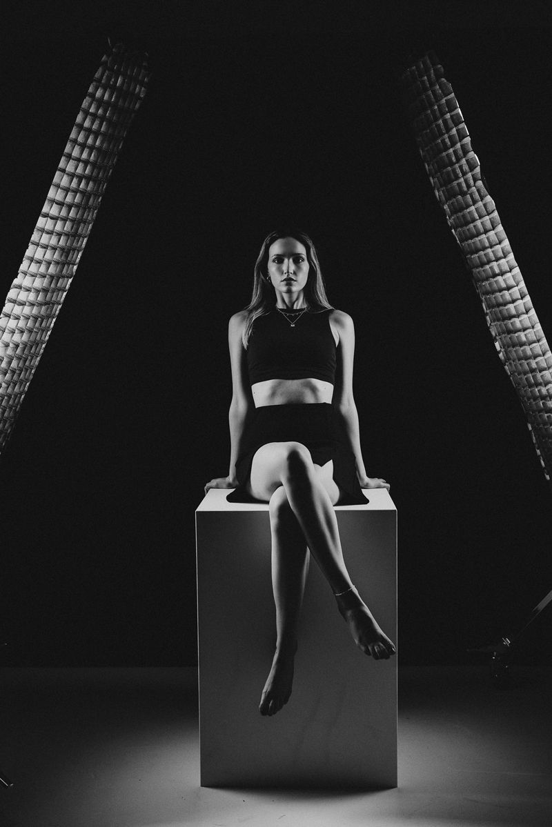 Auf einem Schwarzweißfoto sitzt eine Frau mit übereinandergeschlagenen Beinen auf einem Würfel.