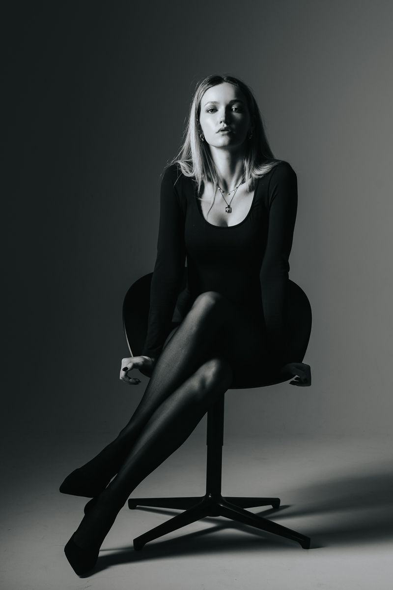 Eine Frau in einem schwarzen Kleid sitzt mit übereinandergeschlagenen Beinen auf einem Stuhl.