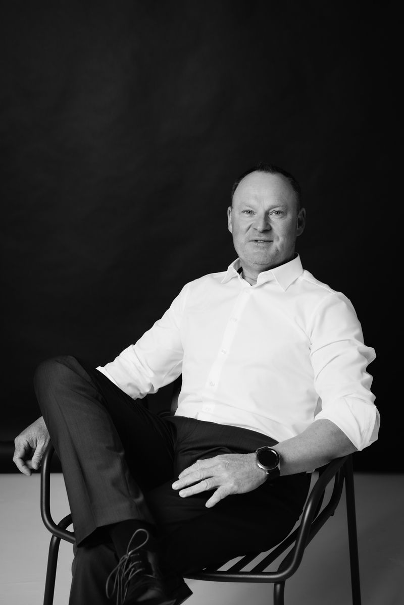 Ein Mann in einem weißen Hemd sitzt mit übereinandergeschlagenen Beinen auf einem Stuhl.