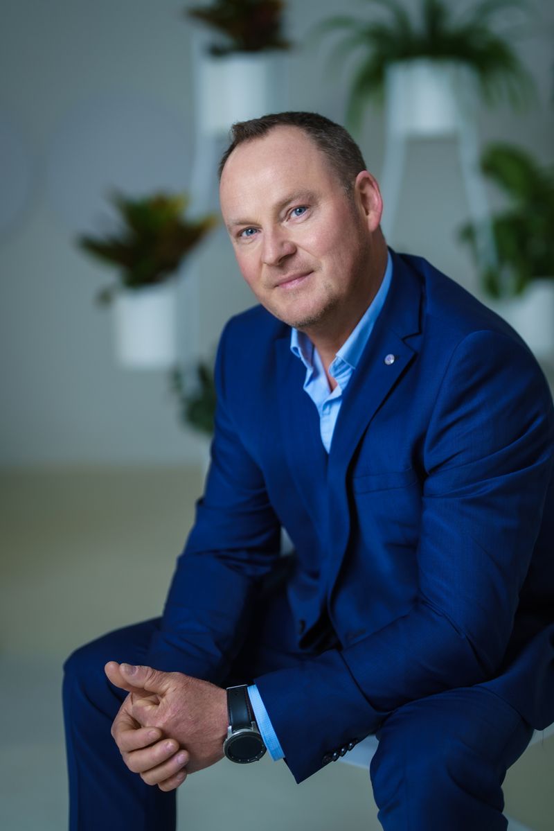 Ein Mann in einem blauen Anzug sitzt mit gefalteten Händen auf einem Hocker.