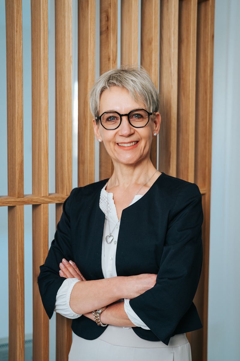 Eine Frau mit Brille steht mit verschränkten Armen vor einem Holzzaun.