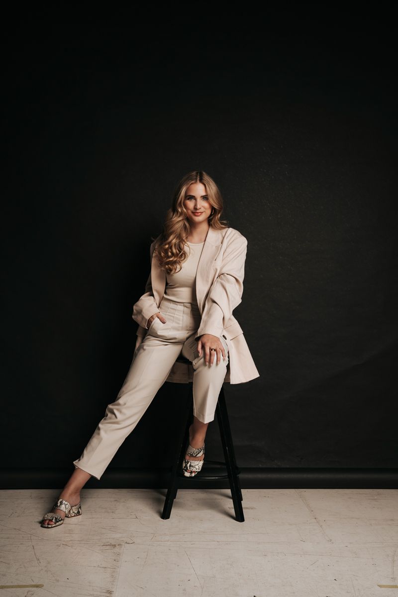 Eine Frau im Anzug sitzt auf einem Hocker vor einem schwarzen Hintergrund.