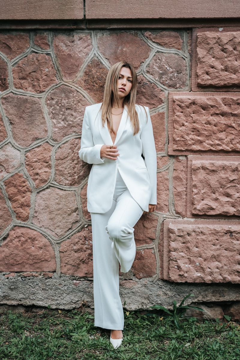 Eine Frau in einem weißen Anzug steht vor einer Backsteinmauer.