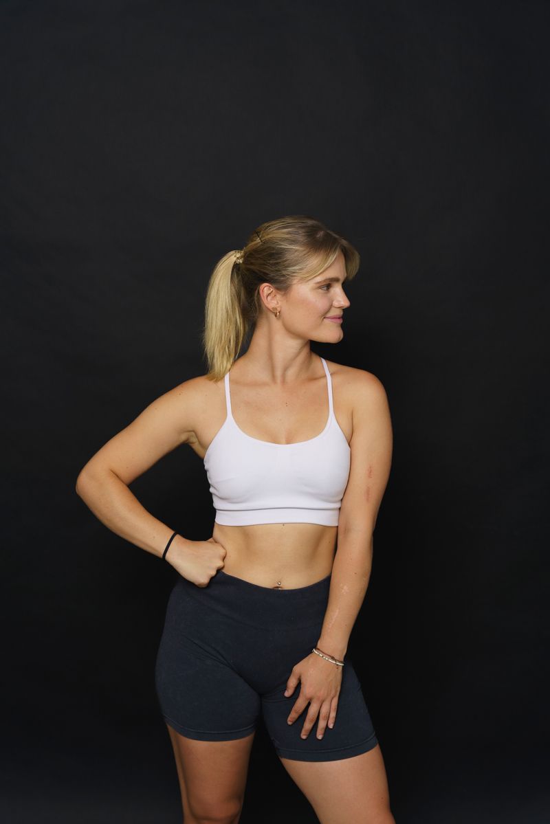Eine Frau in einem weißen Sport-BH und schwarzen Shorts steht vor einem schwarzen Hintergrund.