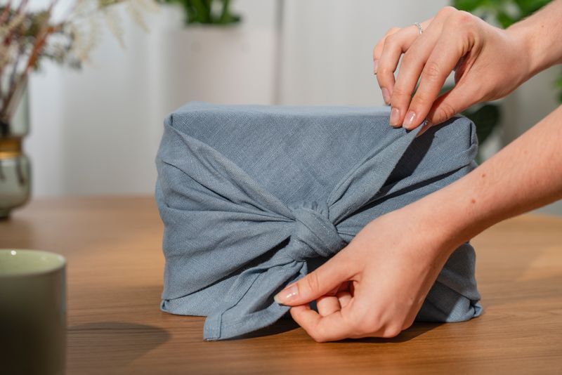 Eine Person verpackt auf einem Holztisch ein Geschenk in ein blaues Tuch.