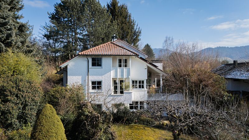 Ein großes weißes Haus mit Ziegeldach ist von Bäumen und Büschen umgeben.