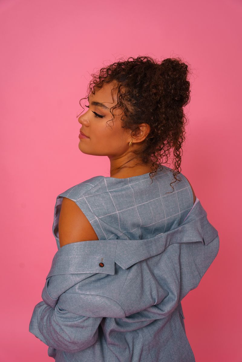 Eine Frau mit lockigem Haar trägt eine blaue Jacke auf rosa Hintergrund.