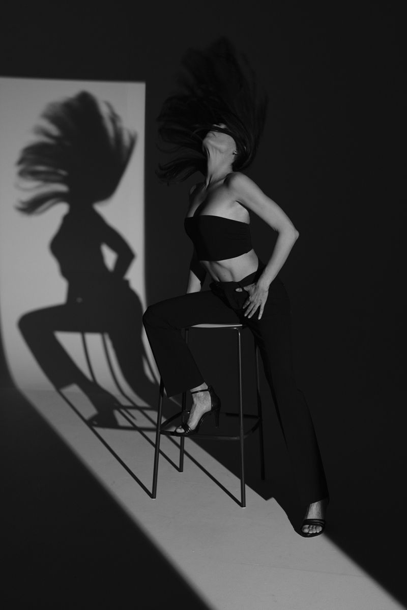 Eine Frau sitzt auf einem Hocker vor einer Wand, auf die ihr Schatten projiziert wird.