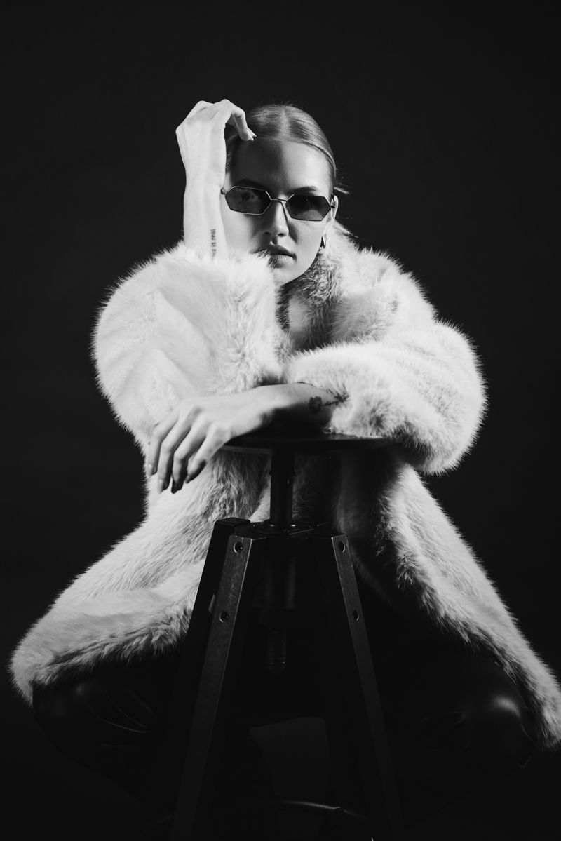 Eine Frau in einem Pelzmantel und mit Sonnenbrille sitzt auf einem Hocker.