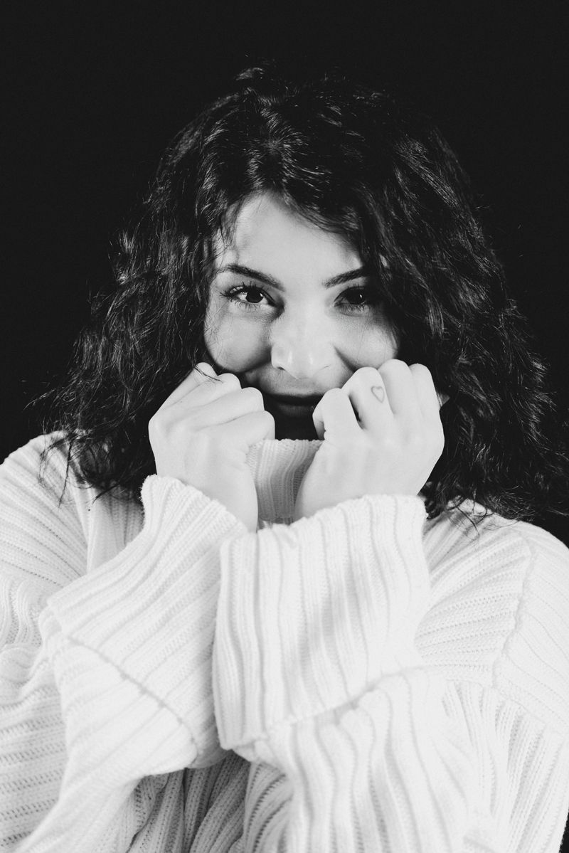 Eine Frau mit lockigem Haar trägt einen weißen Pullover und bedeckt ihr Gesicht mit den Händen.