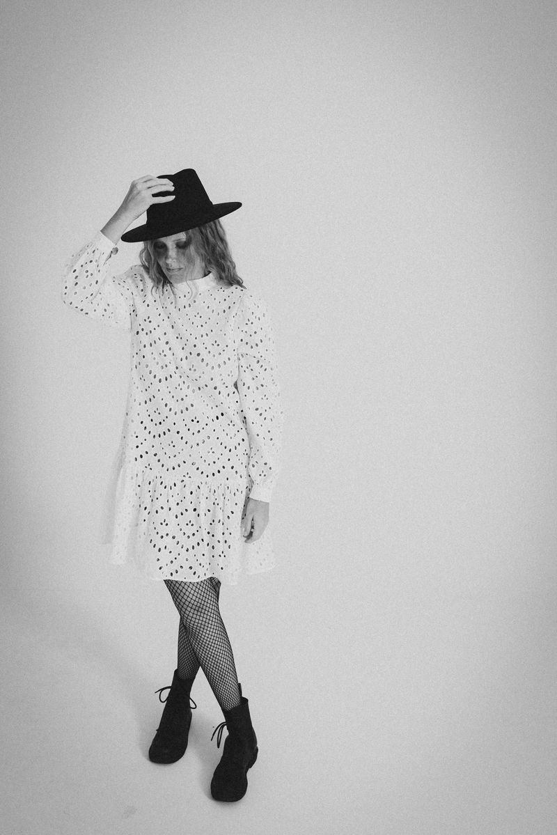 Eine Frau in einem weißen Kleid und einem schwarzen Hut steht vor einer weißen Wand.