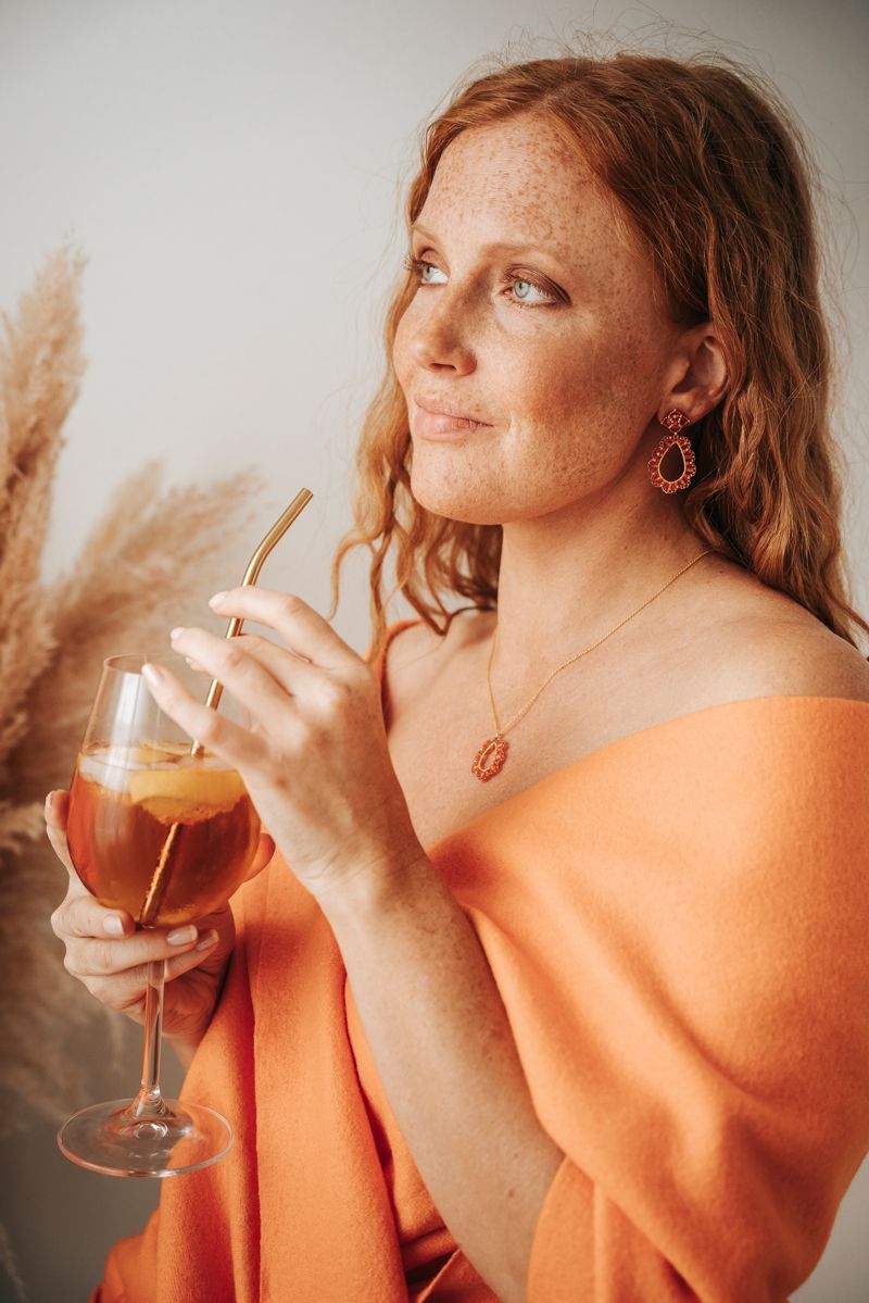 Eine Frau in einem orangefarbenen Kleid hält ein Glas Wein und einen Strohhalm in der Hand.