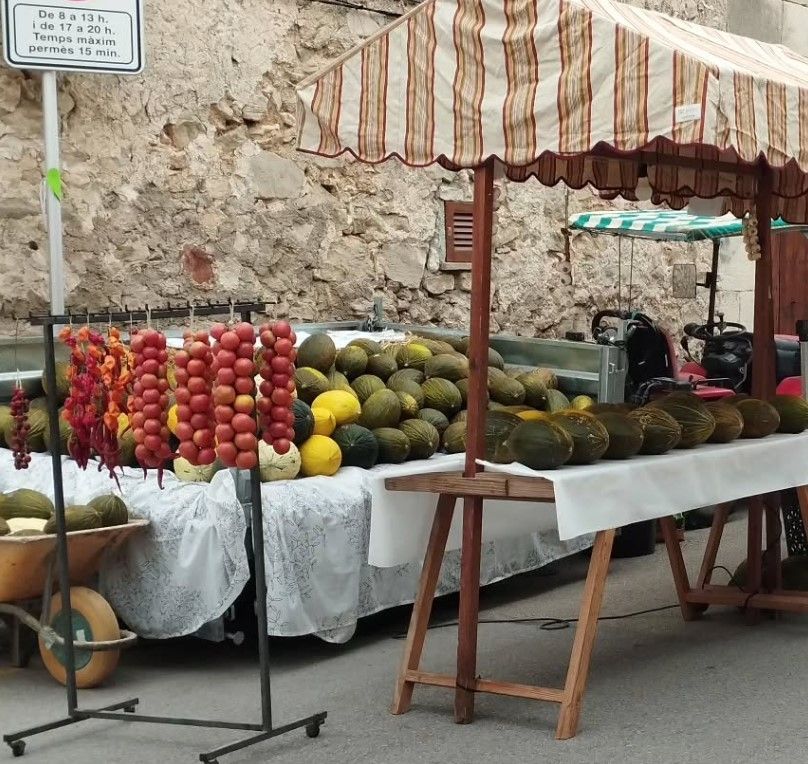 Puesto de mercado con productos, como tomates y melones colgantes, bajo un toldo a rayas. Se ve una carretilla con más fruta.