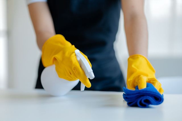Persona que usa guantes amarillos rociando limpiador y limpiando una superficie blanca con un paño azul.