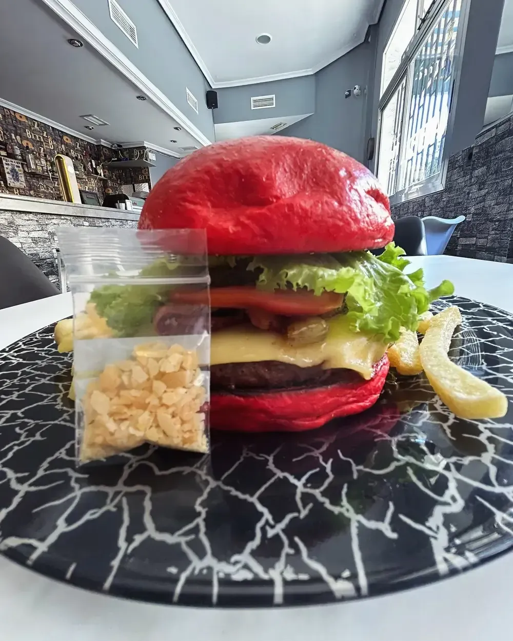 Hamburguesa de pan rojo con queso, lechuga, tomate, pepinillos y patatas fritas en un plato estampado, junto a una bolsa