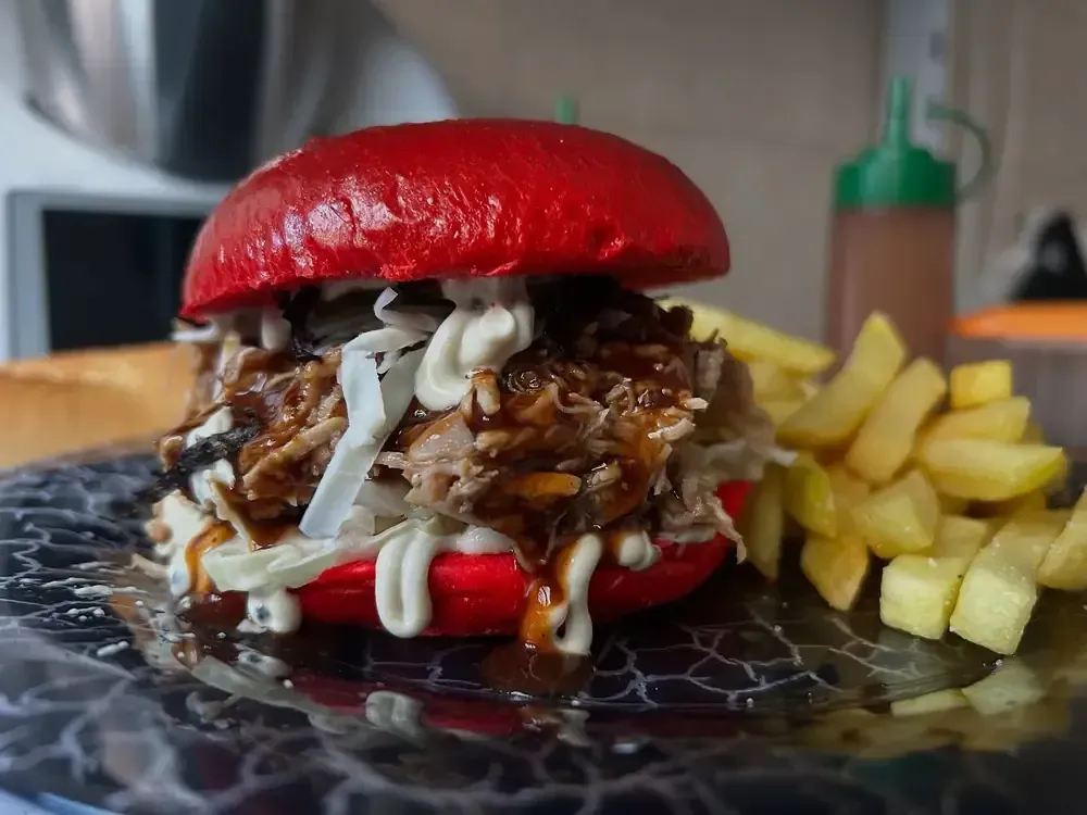 Sándwich de cerdo desmenuzado en pan rojo con ensalada de col, salsa y papas fritas.