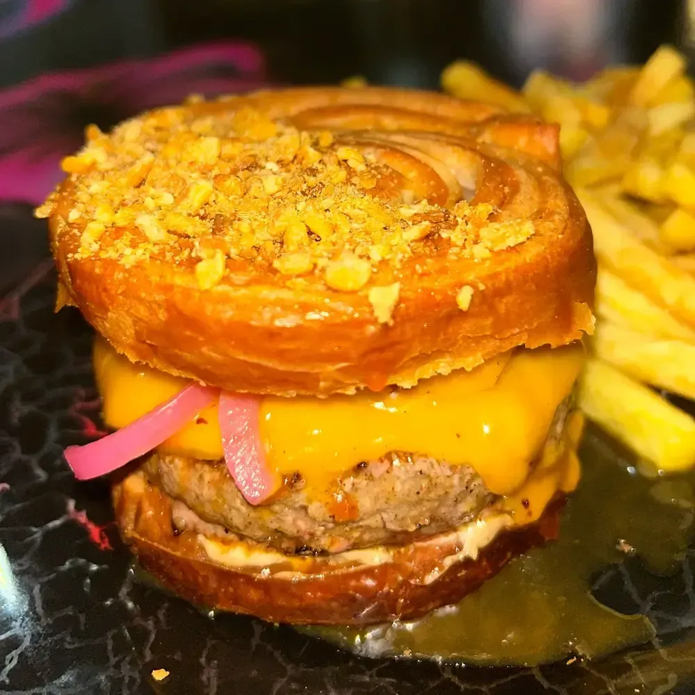 Hamburguesa con pan de pretzel, queso, pepinillos y patatas fritas.