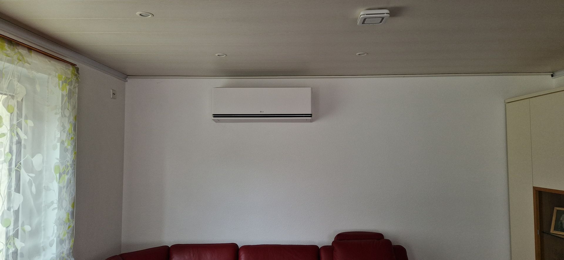 Eine weiße Klimaanlage ist an einer schlichten weißen Wand über einem dunkelroten Sofa in einem Zimmer mit einem durchsichtigen Vorhang angebracht.
