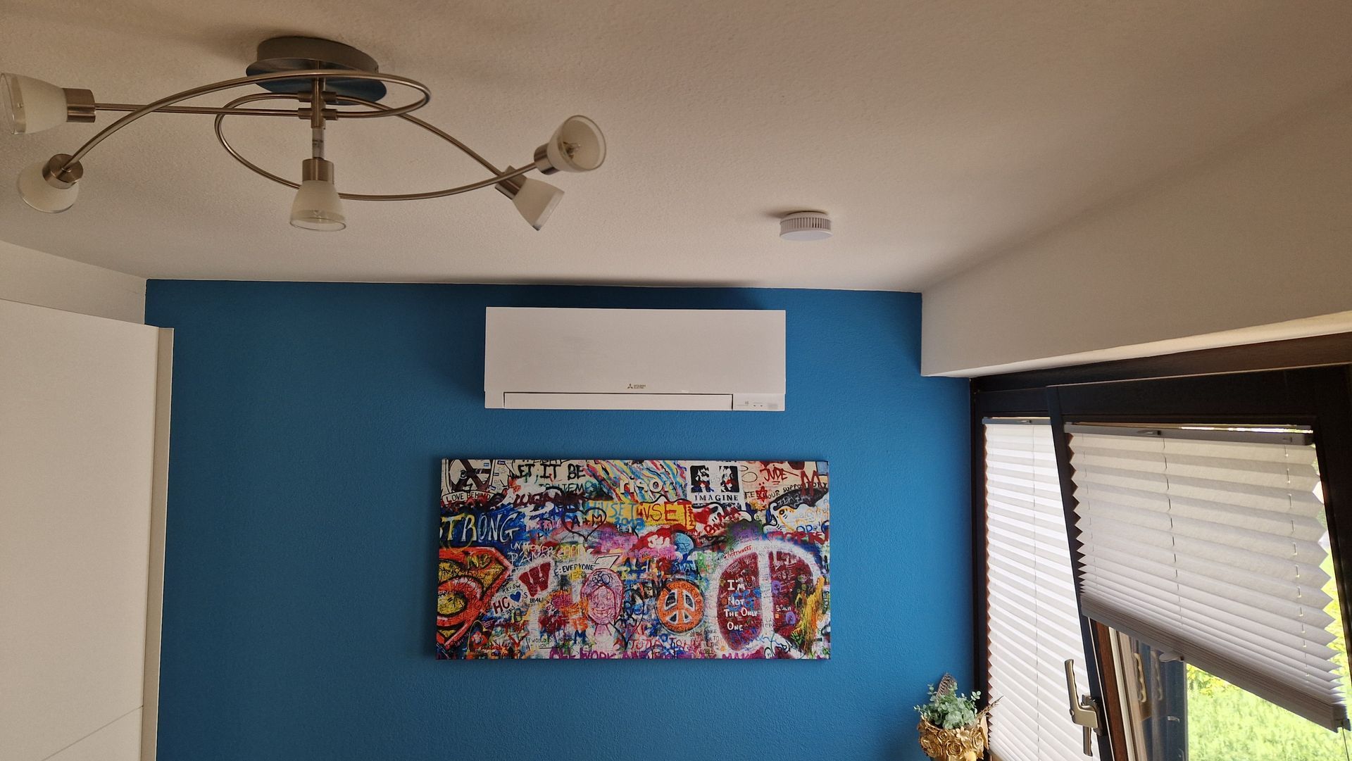 Eine weiße Klimaanlage ist an einer hellblauen Wand über einem farbenfrohen Wandbild aus der Popkultur angebracht.