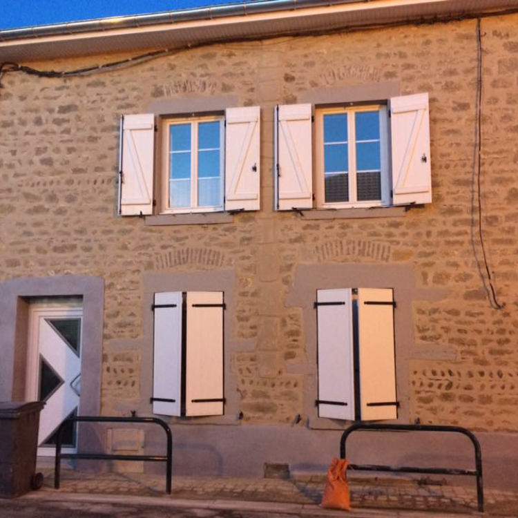 Bâtiment en pierre avec volets blancs, deux fenêtres à l'étage et une porte au rez-de-chaussée.