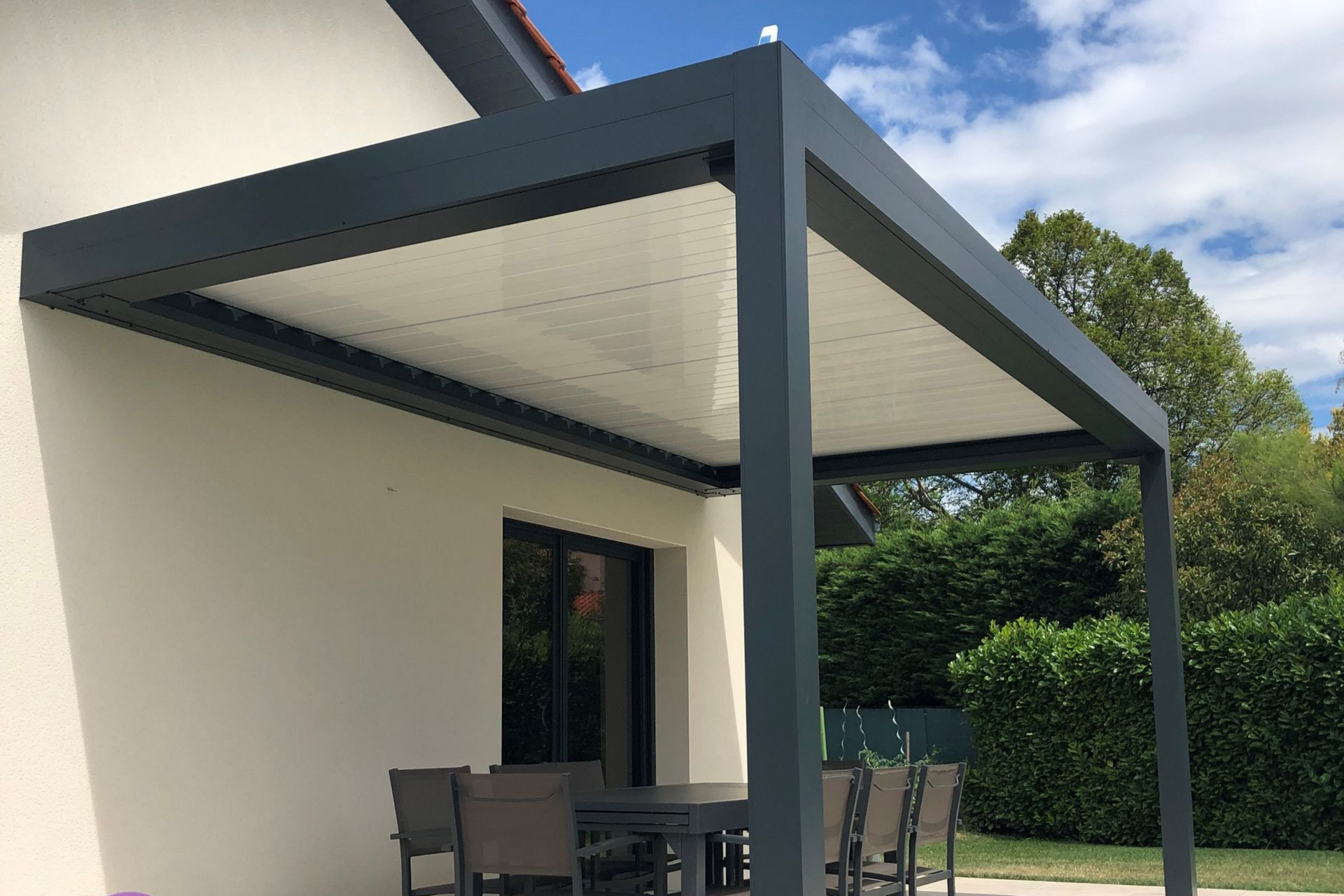 Pergola avec toit rétractable au-dessus d'une table à manger extérieure, structure gris foncé, maison beige clair.