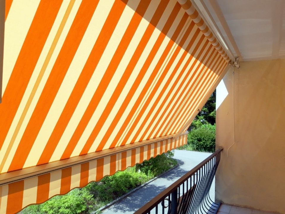 Un store banne à rayures orange et crème s'étendait au-dessus d'un balcon à balustrade noire, donnant sur un chemin et de la verdure.