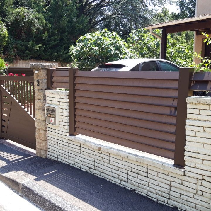 Clôture à claire-voie marron sur un muret en briques, à côté d'un portail marron.