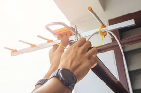 Persona sujetando una antena de TV a un soporte con las manos. Exterior, con luz solar intensa.