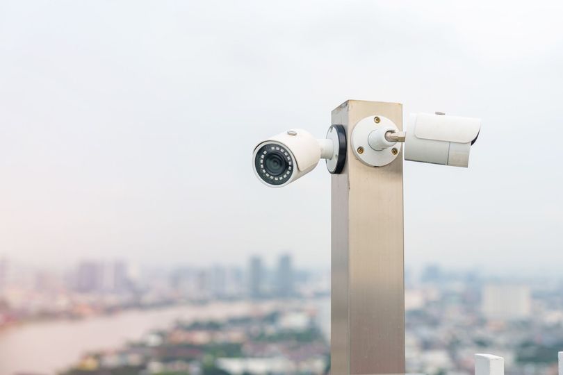 Dos cámaras de seguridad blancas montadas en un poste de metal, con vistas a un paisaje urbano.
