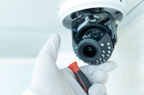 Mano en guante blanco con destornillador ajustando cámara de seguridad.