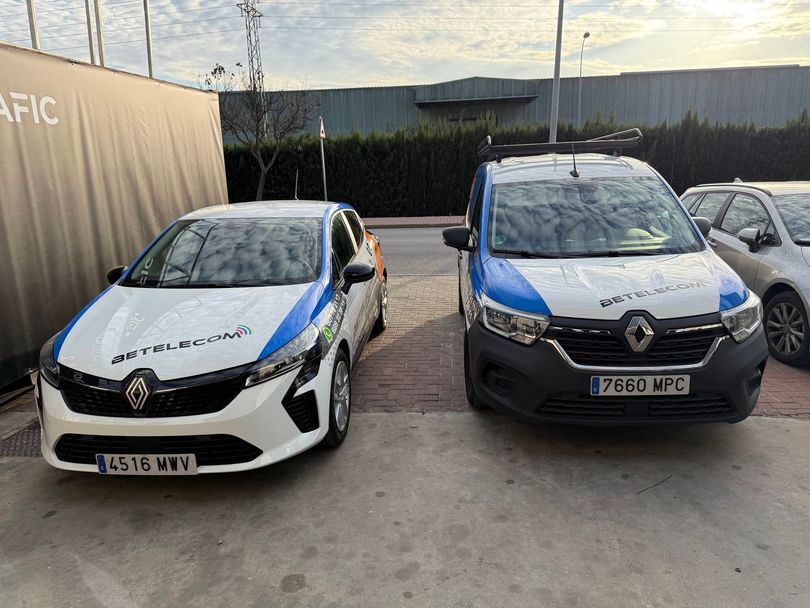 Vehículos con marcas blancas y azules estacionados frente a un edificio. Uno es un coche y el otro una furgoneta, ambos con logotipos.