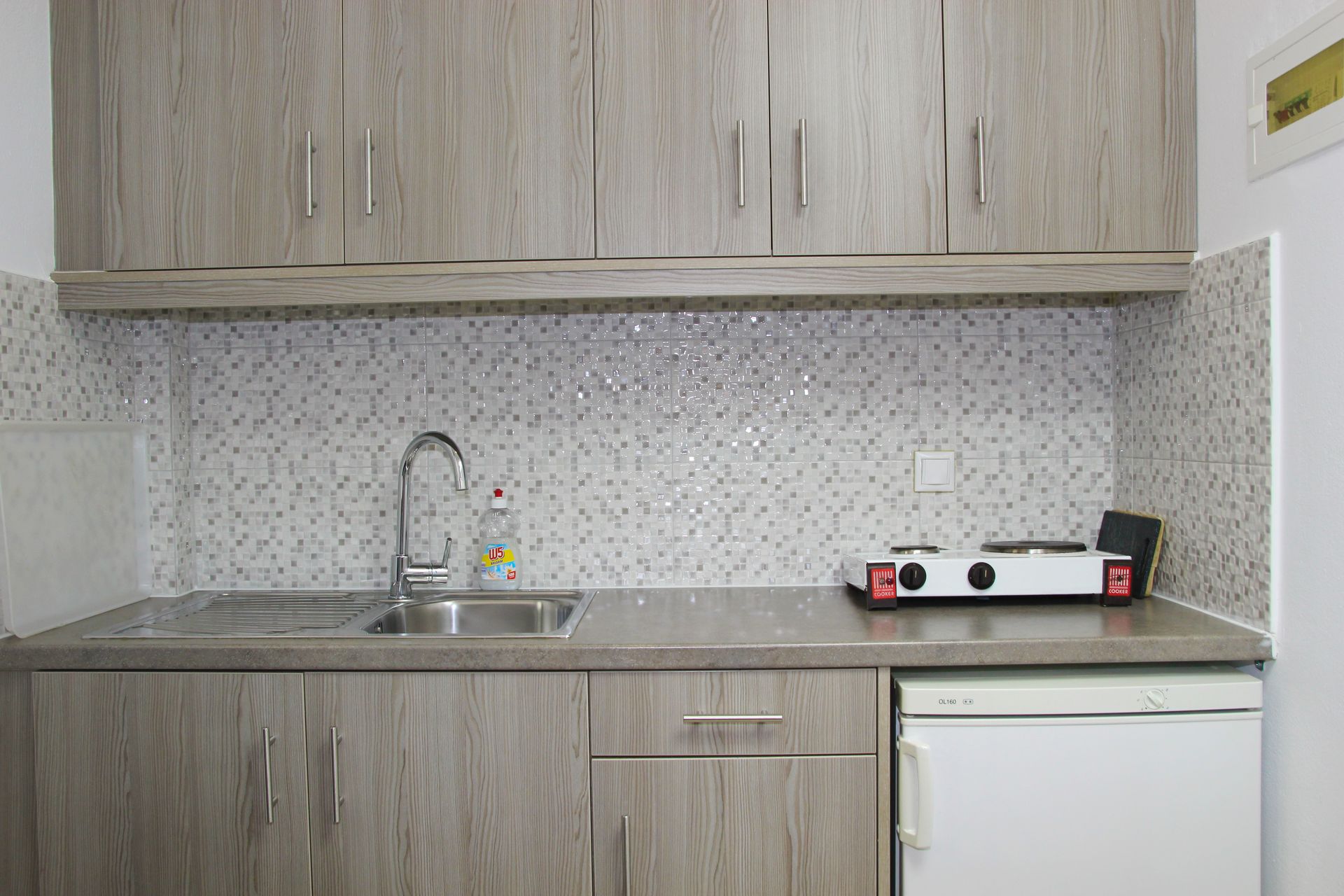 Μικρή κουζίνα με γκρι ντουλάπια, νεροχύτη, φορητή κουζίνα και μίνι ψυγείο.