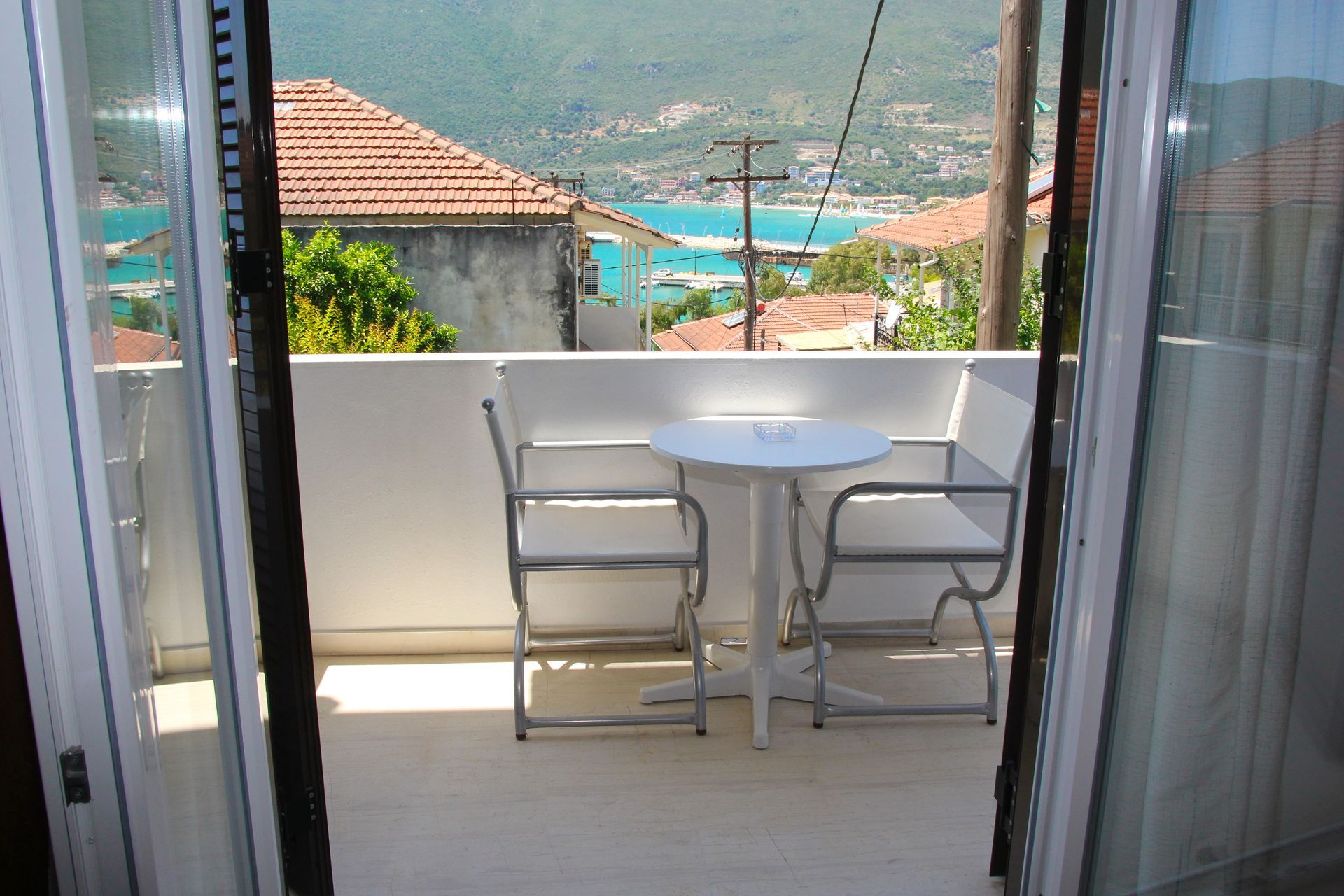 Μπαλκόνι με δύο άσπρες καρέκλες και ένα μικρό τραπέζι με θέα σε έναν μπλε κόλπο και κτίρια με κόκκινες στέγες.