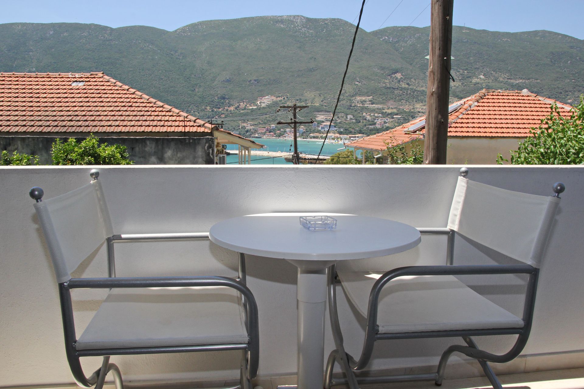 Μπαλκόνι με τραπέζι και καρέκλες με θέα σε έναν κόλπο, λόφους και κτίρια σε μια ηλιόλουστη μέρα.