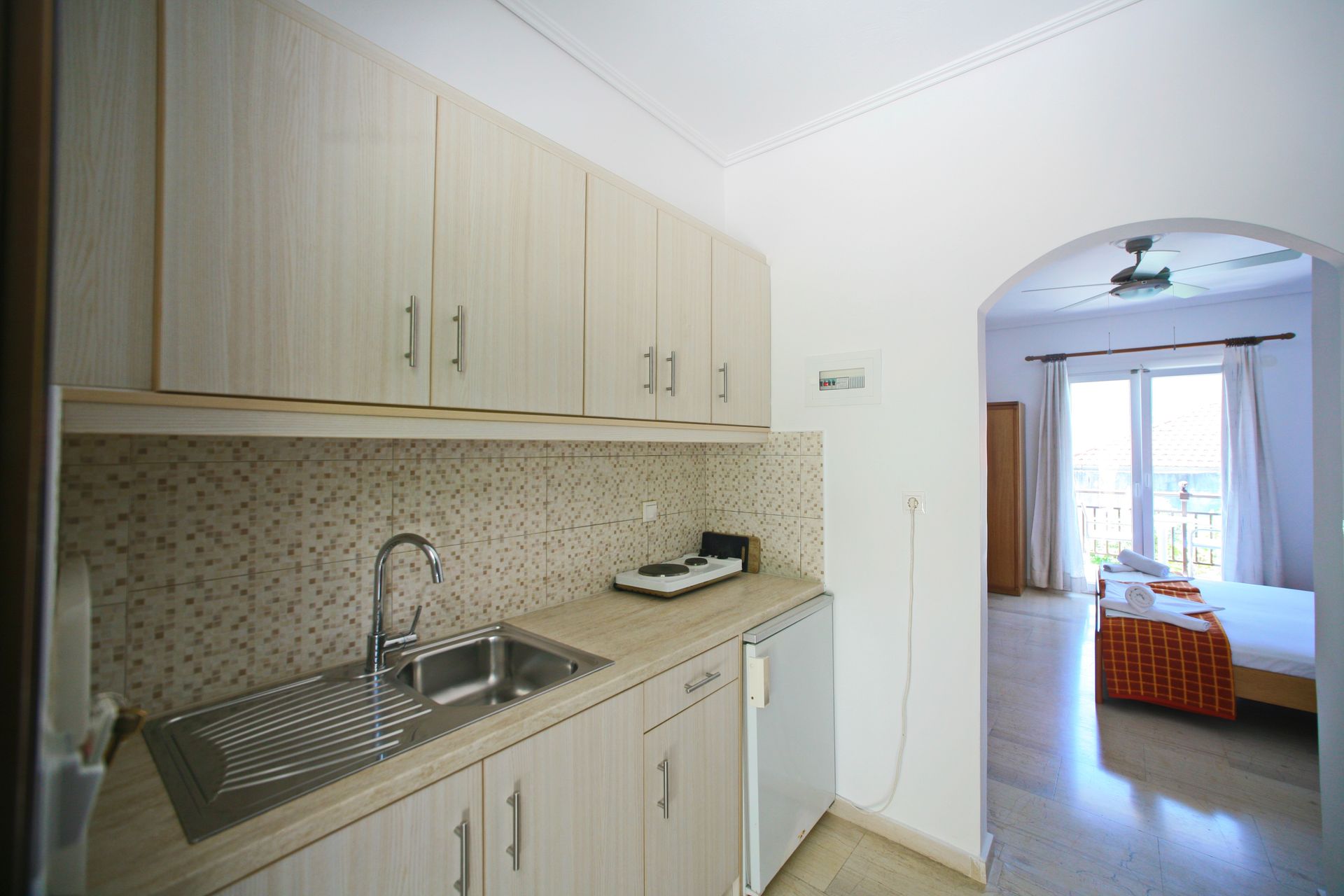 Μικρή κουζίνα με ντουλάπια, νεροχύτη και μικρό ψυγείο. Μια αψίδα οδηγεί σε ένα υπνοδωμάτιο με κρεβάτι και μπαλκόνι.