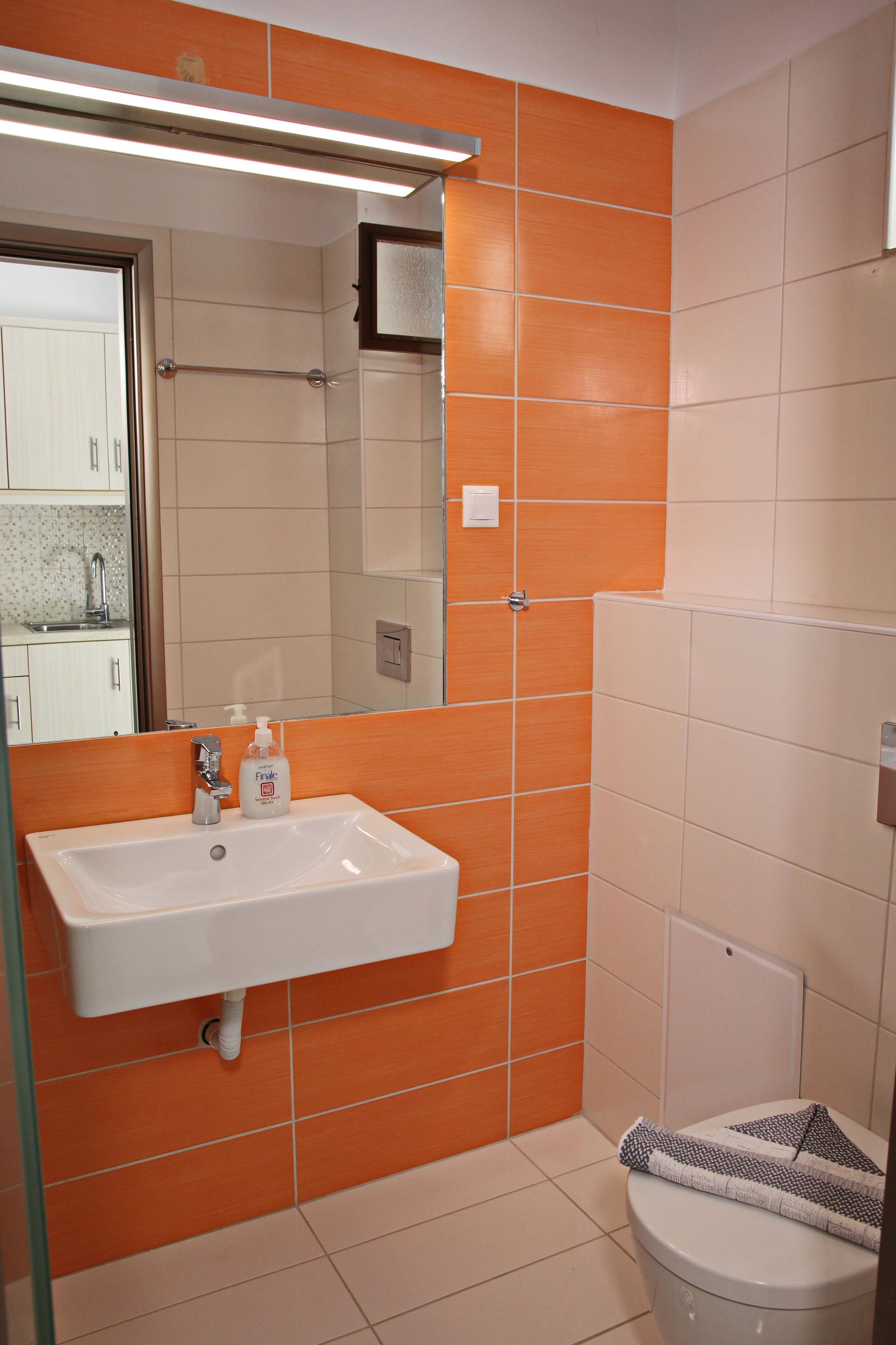 Μπάνιο με πορτοκαλί και κρεμ πλακάκια, λευκό νιπτήρα, καθρέφτη και τουαλέτα.