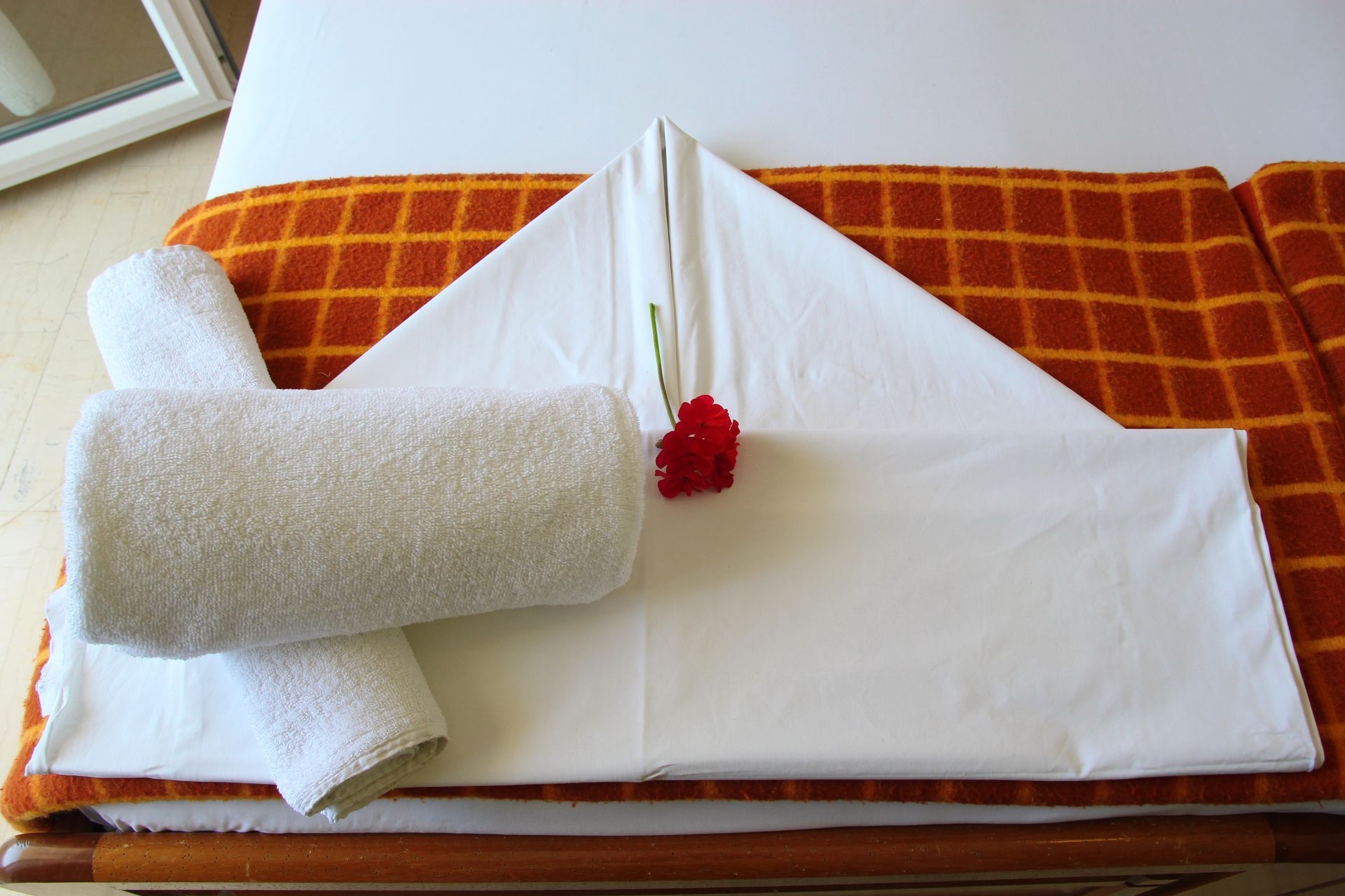 Λευκές πετσέτες τοποθετημένες σε ένα κρεβάτι, με διπλωμένο σχήμα και τυλιγμένες πετσέτες, με ένα κόκκινο λουλούδι στην κορυφή.