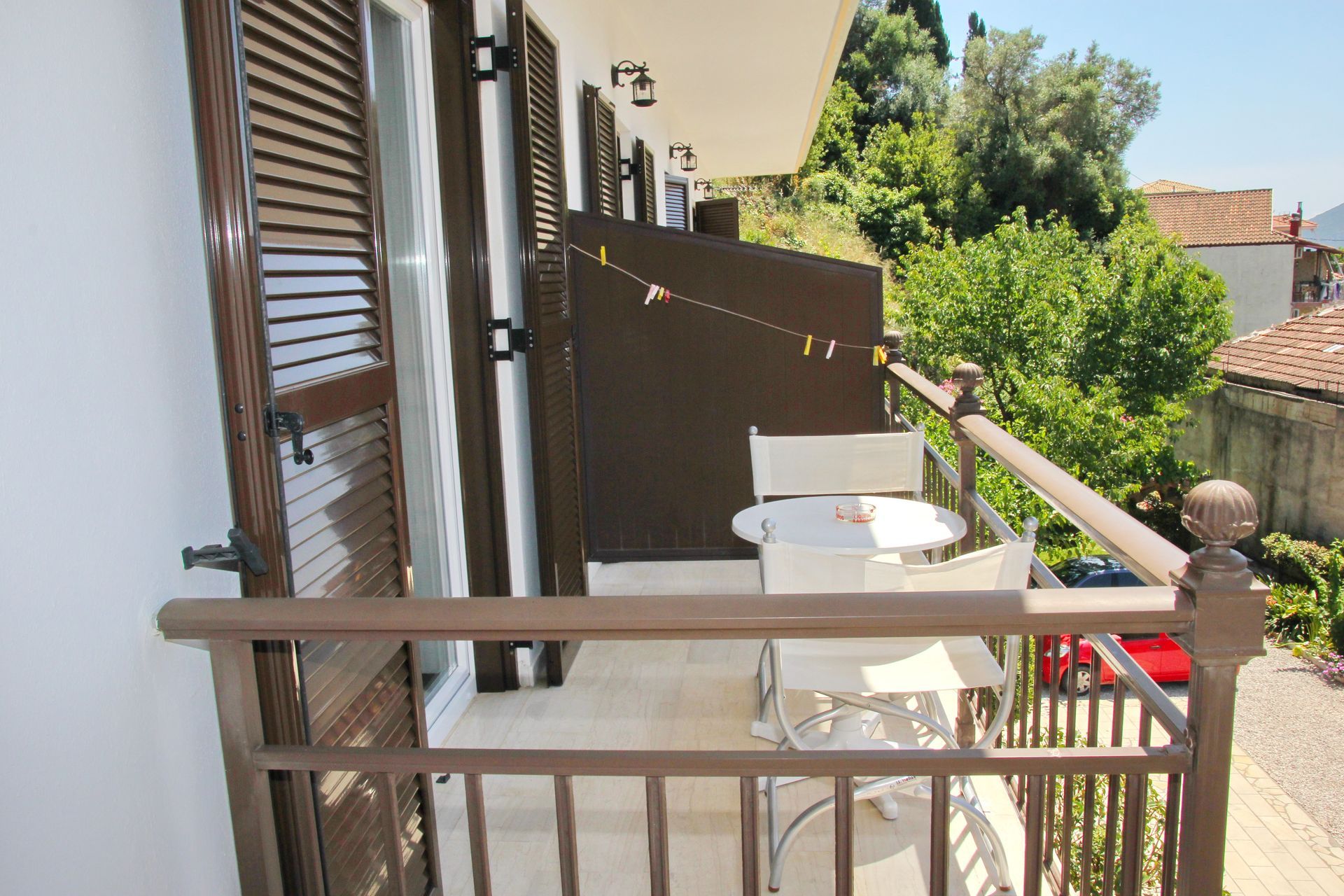 Μπαλκόνι με λευκά έπιπλα, καφέ κιγκλίδωμα και παντζούρια, με θέα σε δέντρα και κτίρια.