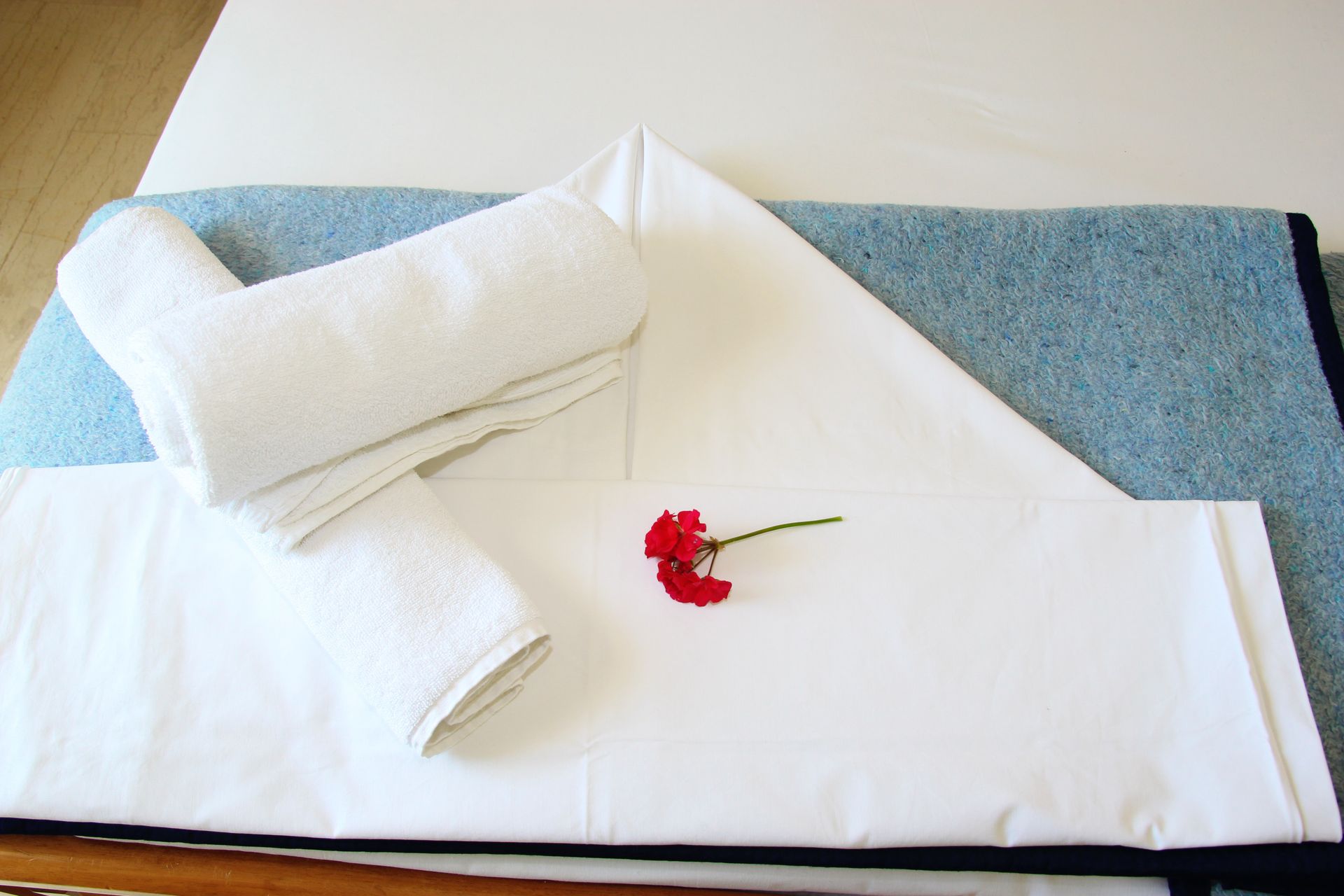 Διπλωμένες λευκές πετσέτες, ένα λευκό σεντόνι με διπλωμένη γωνία και ένα κόκκινο λουλούδι σε ένα μπλε χαλάκι.