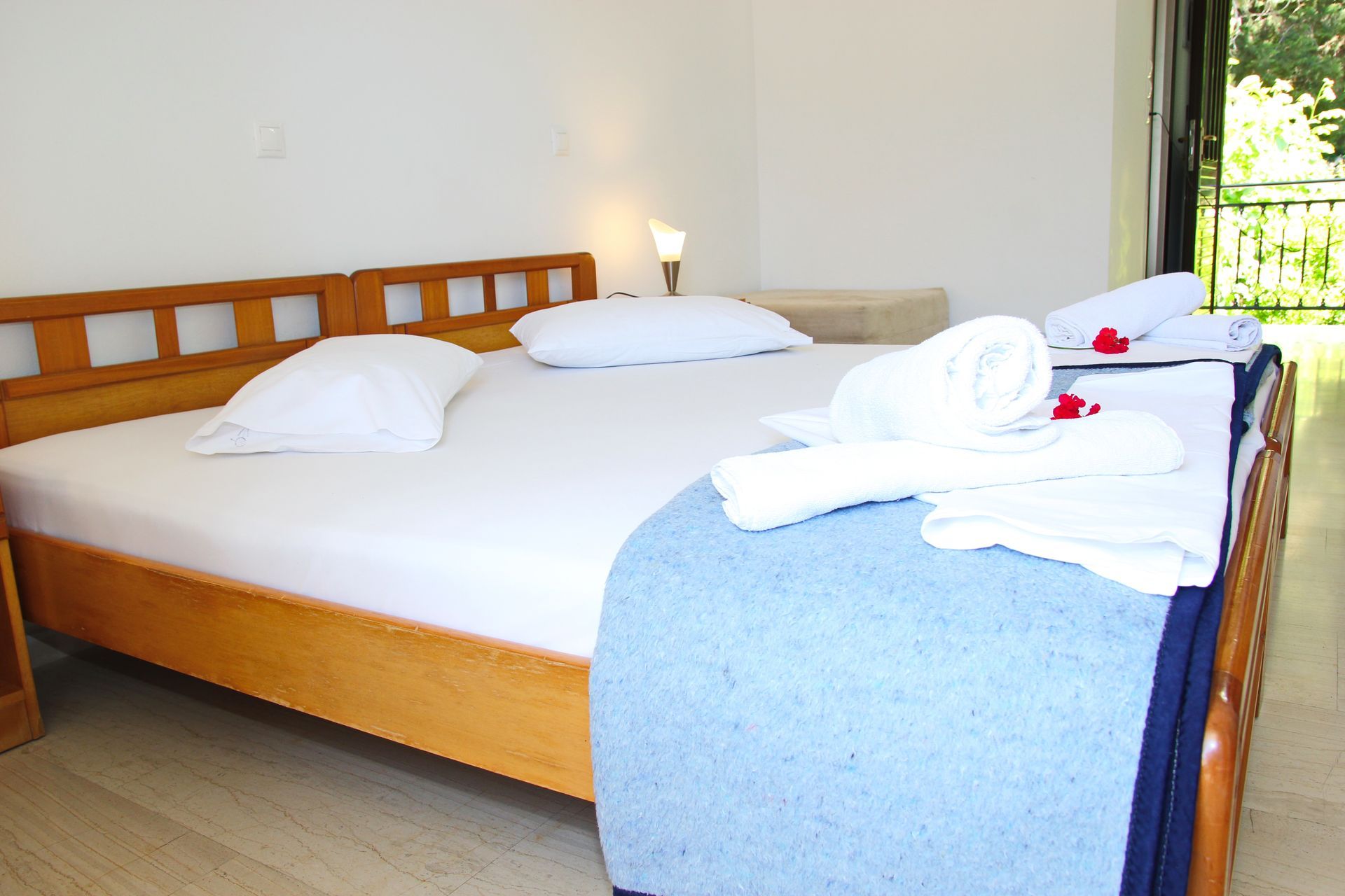 Υπνοδωμάτιο με ανοιχτόχρωμο ξύλινο σκελετό κρεβατιού, λευκά κλινοσκεπάσματα, μπλε κουβέρτα και ρολά πετσετών. Ορατό μπαλκόνι.