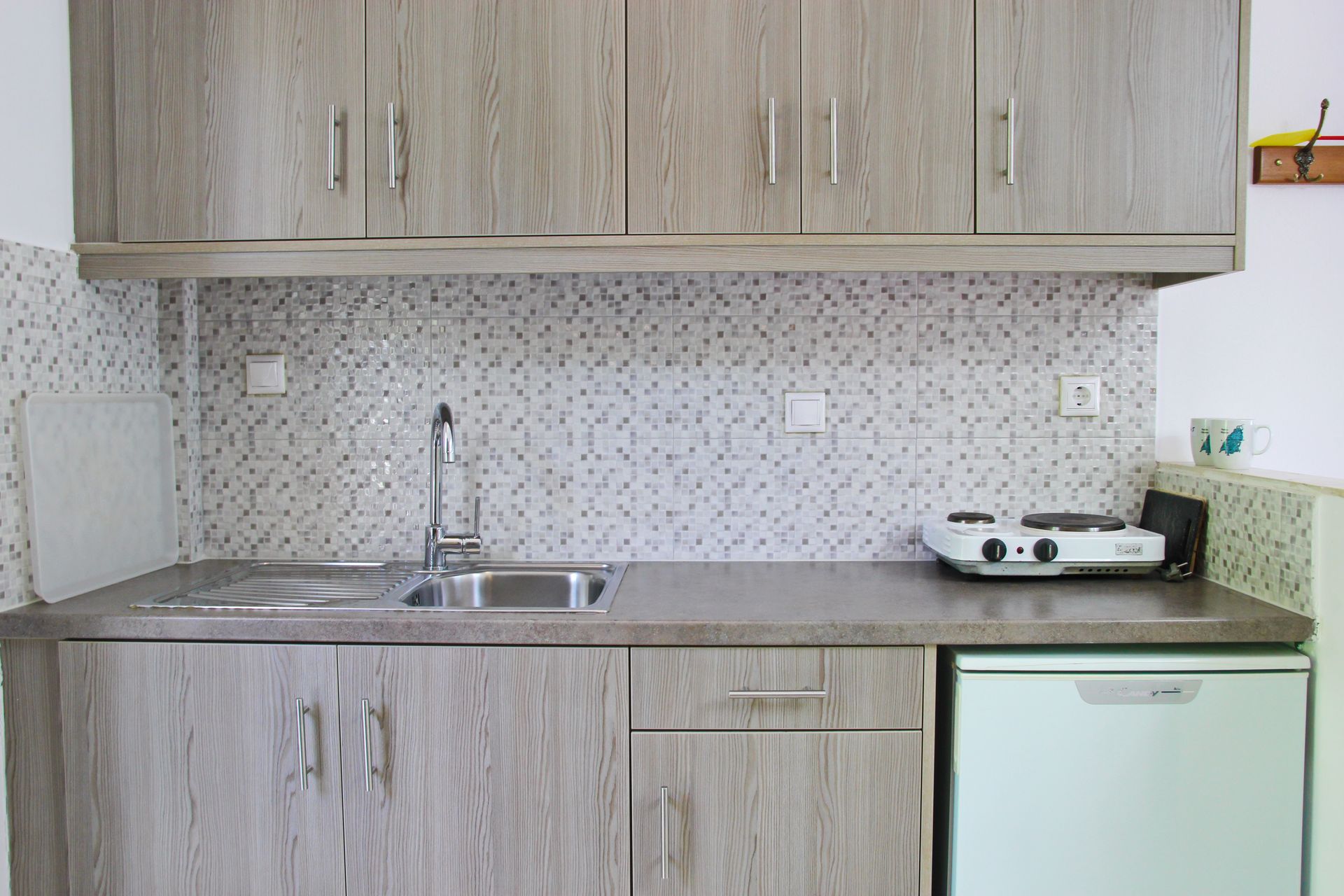 Κουζίνα με ξύλινα ντουλάπια, νεροχύτη, εστία μαγειρέματος και μικρό ψυγείο. Λευκή πλακόστρωση.