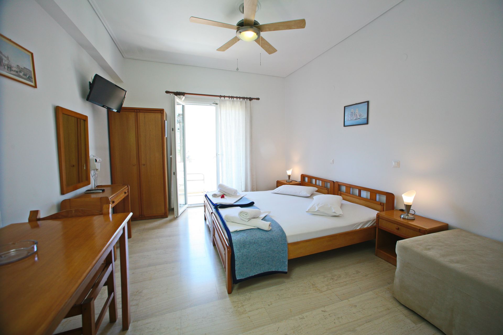 Φωτεινό υπνοδωμάτιο με διπλό κρεβάτι, ξύλινα έπιπλα και μπαλκονόπορτα. Διακρίνεται ένας ανεμιστήρας οροφής.