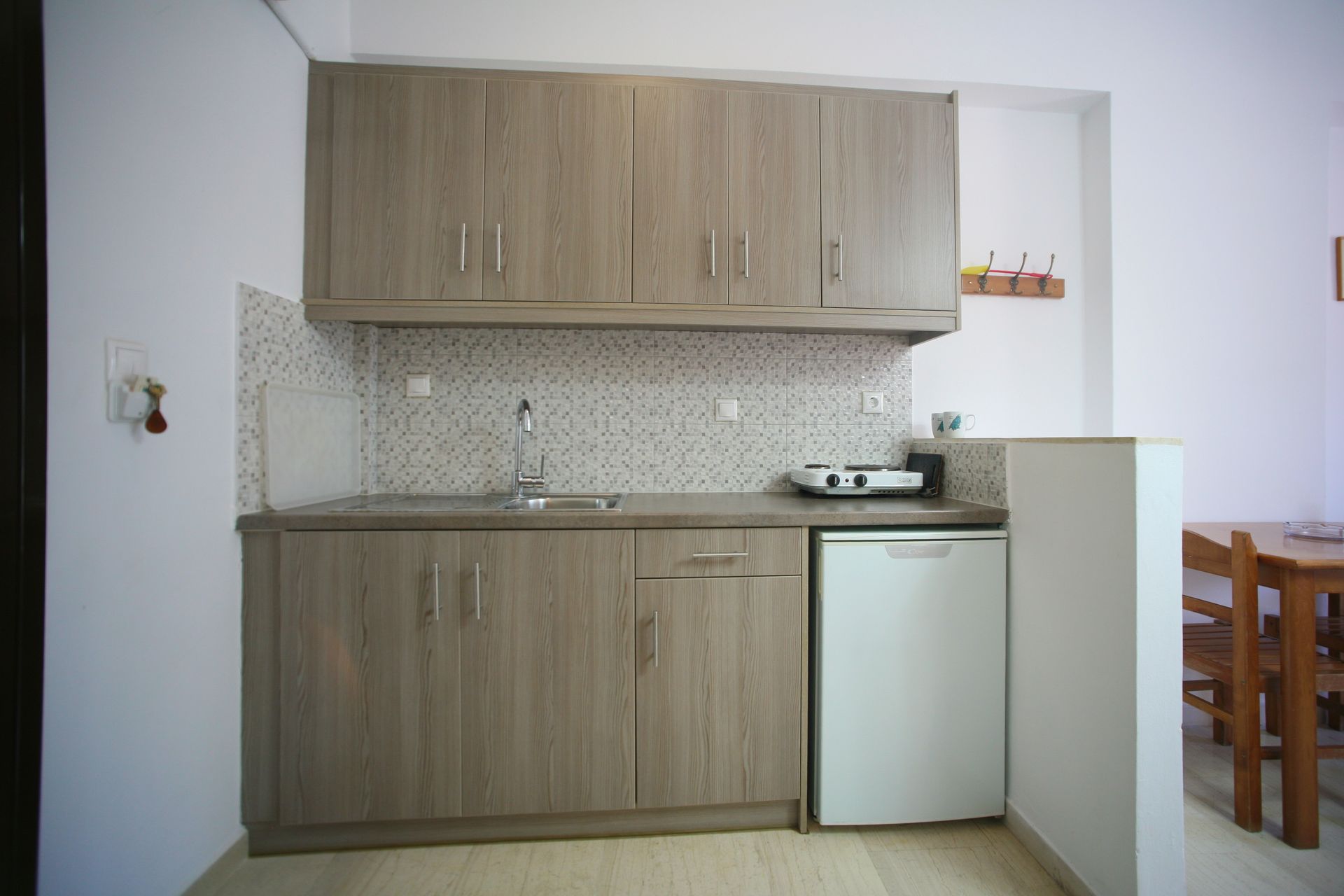 Μικρή κουζίνα με ανοιχτόχρωμα ξύλινα ντουλάπια, πάγκο και μίνι ψυγείο.