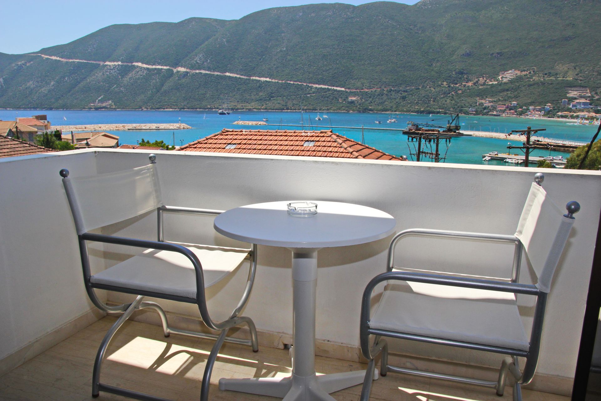 Μπαλκόνι με δύο λευκές καρέκλες και ένα τραπέζι με θέα σε έναν κόλπο και βουνά.