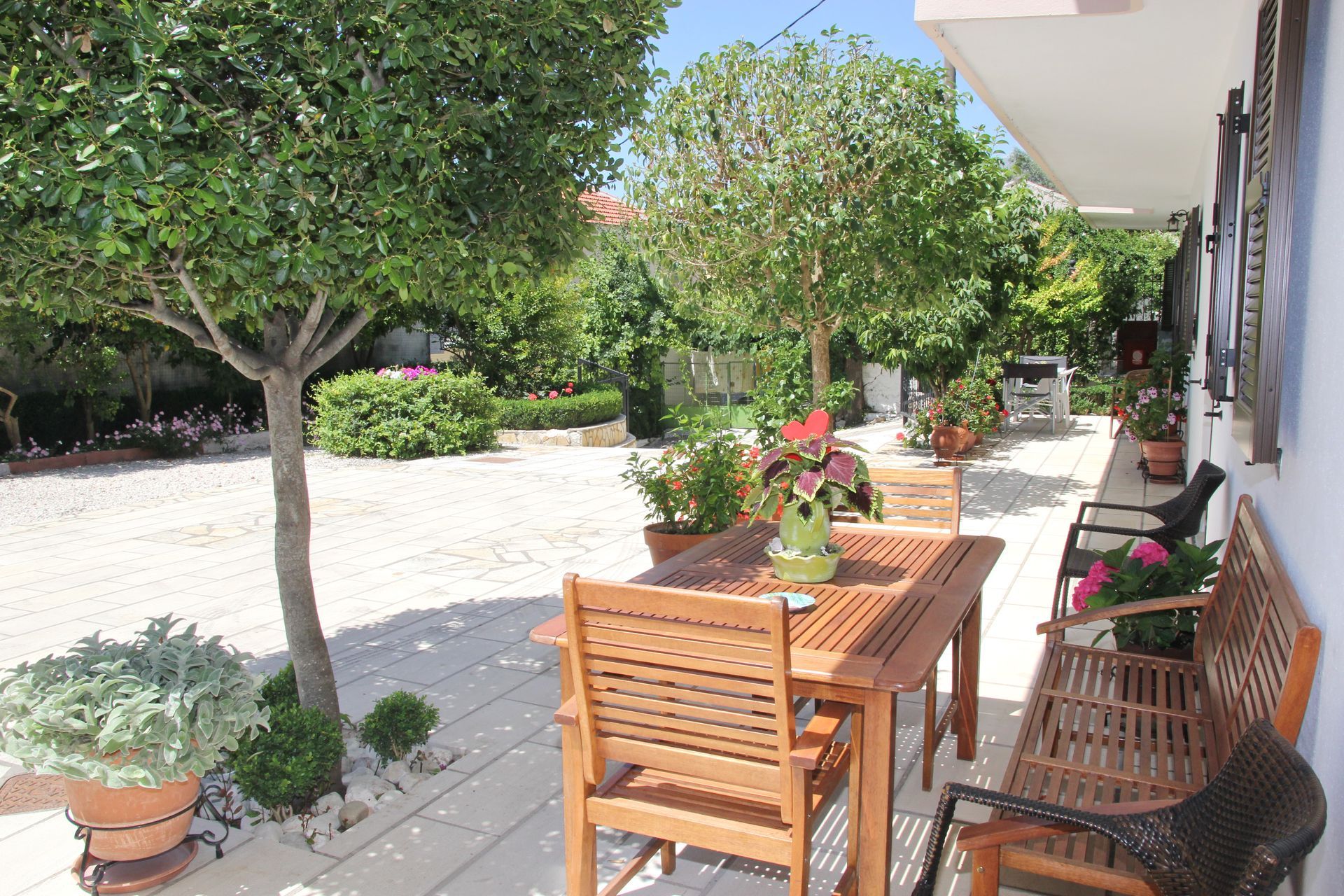 Ξύλινα έπιπλα βεράντας σε βοτσαλωτή περιοχή, περιτριγυρισμένα από δέντρα και φυτά σε γλάστρες, σε ηλιόλουστο εξωτερικό περιβάλλον.