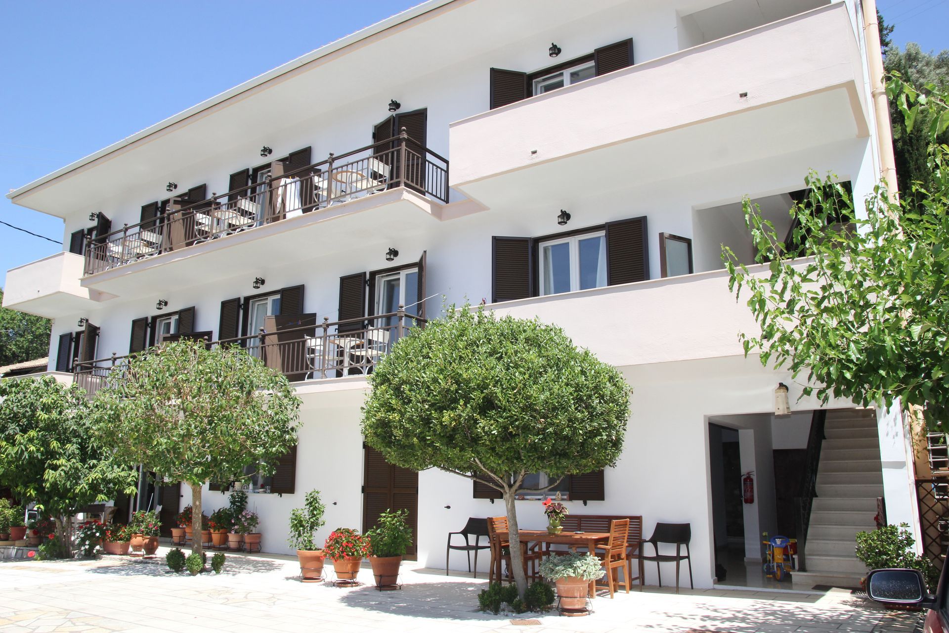 Λευκό πολυώροφο κτίριο με μπαλκόνια, καφέ παντζούρια και γλάστρες με φυτά.