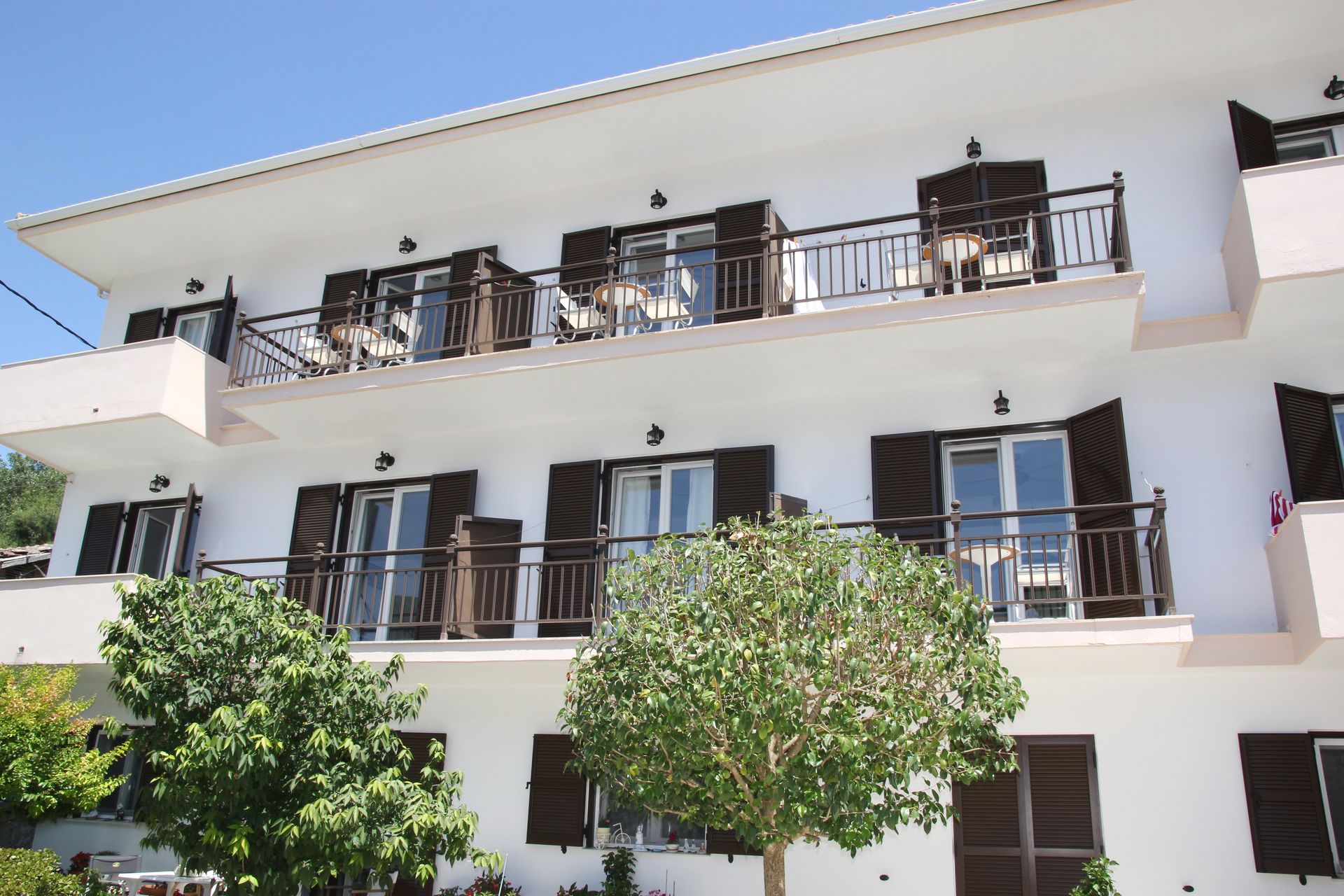 Λευκό πολυώροφο κτίριο με μπαλκόνια, καφέ παντζούρια και δέντρα.